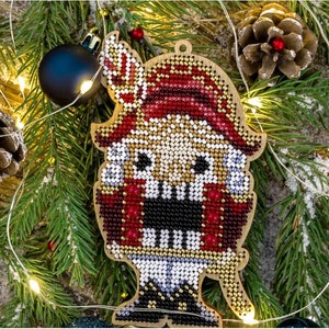 May include: Wooden Christmas ornament in the shape of a nutcracker. The ornament is decorated with red, white, black, and gold beads. The background includes pine needles, a pine cone, and a dark blue ornament.