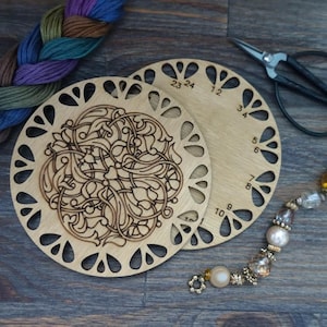 May include: Two wooden thread organizers with laser-cut designs and numbers, alongside embroidery floss and scissors. One organizer features an intricate floral pattern, while the other has numbers around the edge. A beaded charm hangs nearby.