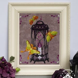 May include: A framed cross-stitch embroidery on a mottled gray fabric featuring a black lantern with a glowing purple candle inside, surrounded by three yellow and green moths, with small purple floral accents and a cream-colored frame.