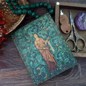 May include: An antique-style journal with a woman illustration, green and blue floral patterns, and a wooden box containing scissors, a wooden tool, and a purple stone. Also, a gold tray with beads and a strand of amber beads.
