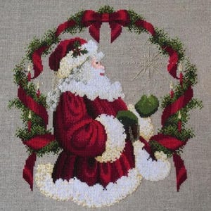 May include: Cross-stitch depiction of Santa Claus in a red suit with white trim, holding a green mitten. He is surrounded by a wreath of greenery, red ribbons, and candles. A large red bow sits at the top of the wreath.