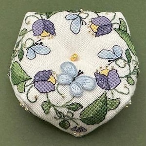 May include: A white, pentagon-shaped pincushion with embroidered floral and butterfly designs. The flowers are purple and yellow, and the butterflies are light blue. Green leaves and gold beads accent the design. A gold bead is in the center.