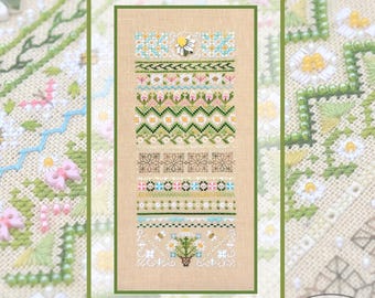 May include: A framed cross-stitch sampler featuring various floral and geometric patterns in shades of green, pink, blue, and brown. The sampler includes a daisy embellishment and is set against a neutral background.