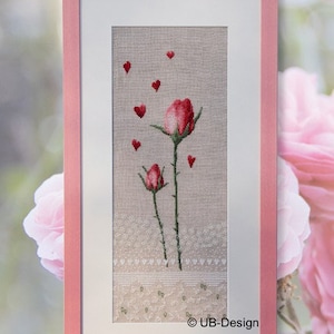 Puede incluir: Un bordado de punto de cruz enmarcado que presenta dos rosas rosas con tallos y hojas verdes. Las rosas están rodeadas de pequeños corazones rojos sobre un fondo de lino beige. La parte inferior del bordado tiene un diseño de encaje blanco y el texto "UB-Design" está en la esquina inferior derecha.