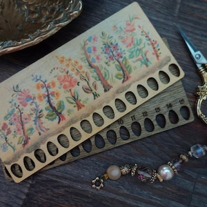 May include: A wooden needle gauge with floral artwork and oval holes, paired with a darker wooden gauge. Gold scissors and a beaded bracelet are also present. The items are arranged on a dark wood surface.