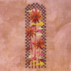 May include: A cross-stitch embroidery on mottled pink fabric featuring three stylized orange and red flowers with yellow cloud-like shapes. The floral design is framed by a dark brown checkered border with a rounded top edge.