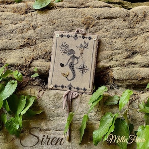 May include: A framed cross-stitch featuring a mermaid design, holding a heart and a compass. The mermaid is black, with a beige background and a decorative border. The word "Siren" is at the bottom left, and "Mille Fleur" is at the bottom right.