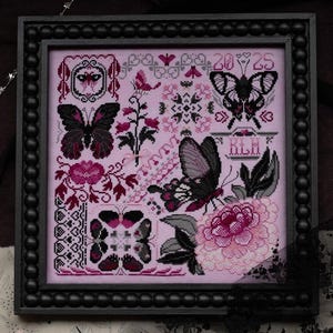 May include: A square cross-stitch sampler in a black beaded frame featuring a light pink fabric background with black, dark pink, and white thread designs of butterflies, flowers, and the text 2025 and RLH in a decorative border style.