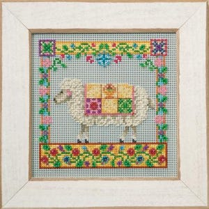 May include: A framed cross-stitch embroidery featuring a white sheep standing in profile. The sheep wears a colorful patchwork blanket with yellow, pink, and green squares. The design is bordered by a floral pattern in pink, green, and blue.
