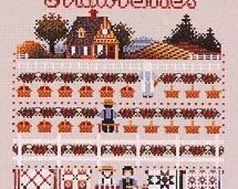 May include: A cross-stitch embroidery pattern on light fabric featuring the text Strawberries Forever. The design depicts a house, strawberry fields with baskets, and two figures standing at a table labeled Pies N Jam, surrounded by various quilt patterns.