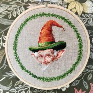 May include: A cross-stitch embroidery in a wooden hoop features a whimsical gnome with an orange hat and green brim. The gnome is surrounded by a green leaf border. The background is a neutral fabric.