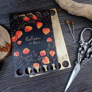 Könnte beinhalten: Eine Holzgarn Schale mit 10 Löchern zum Aufbewahren von Garn. Die Schale ist mit einem schwarzen und orangefarbenen Herbstlaubdesign und dem Text "Autumn is here" verziert.