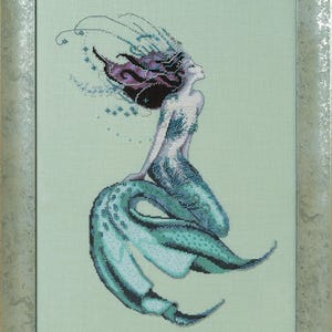 May include: Cross-stitch artwork of a mermaid with flowing teal and purple hair, a teal tail, and a silver frame. The mermaid is depicted in a graceful pose, with her head tilted upwards. The background is a soft, light green colour.
