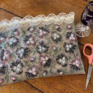 May include: A beige floral embroidered pouch with a scalloped lace trim. The pouch features a pattern of dark and light pink flowers with green stems and leaves. A purple glass and pink scissors with a gold tassel are also present.