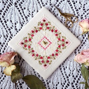 May include: A square, off-white cross-stitch embroidery with a floral design in red and green. The design is centered with a pink and gold heart. Gold scissors rest on the corner, with dried roses nearby. The embroidery is on a white, lace-like background.