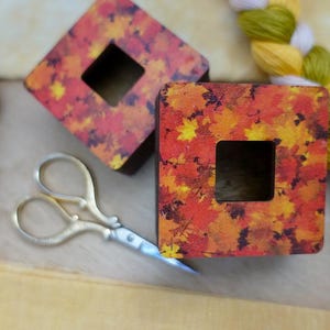 May include: Two square wooden boxes with a fall leaf design and a square hole in the center. The boxes are on a wooden surface with a pair of gold scissors.
