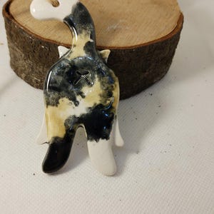 May include: A ceramic cat ornament with a glossy finish. The cat is white, black, and tan with a cross-shaped cut-out. It has a jute twine loop for hanging. The ornament is placed on a wooden slice.