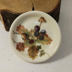 May include: A small, cream-coloured ceramic dish with a detailed botanical design. The illustration features an acorn, mushrooms, berries, and leaves in shades of brown, green, and purple. The dish has a slightly cracked appearance, adding to its vintage charm.