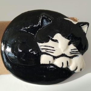 May include: A circular ceramic brooch featuring a glossy black and white cat curled into a sleeping position. The cat has a black body and tail with a white face, chest, and paws, detailed with black painted whiskers and closed eyes.