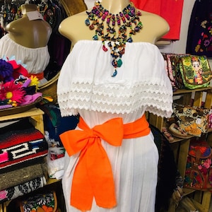 May include: A white, strapless dress with a lace trim and an orange sash tied in a bow. A colorful beaded necklace adorns the mannequin. The dress is displayed in a shop with other colorful items.
