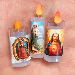 May include: Three miniature candles with religious imagery. Each candle has a clear plastic base and a printed paper wrap depicting religious figures. The flames are orange and yellow. The candles are held in a hand.