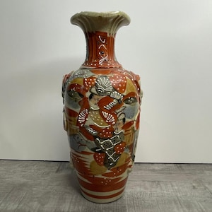 May include: A tall, ceramic vase with a flared rim and a wide body. The vase features a detailed, hand-painted design with figures in traditional clothing, in shades of red, orange, and gray. The vase has a textured surface and a cream-colored base.