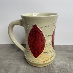 May include: A ceramic mug with a cream-colored glaze and red leaf designs. The mug has a handle and a rounded bottom.