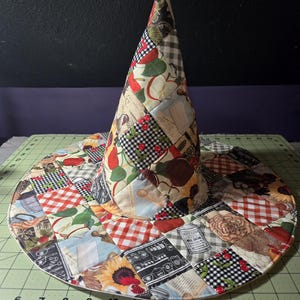 May include: A conical hat constructed from a patchwork of fabric squares featuring various patterns including red apples, black and white gingham, sunflowers, and kitchen-themed illustrations. The hat sits on a green cutting mat with visible inch markings.