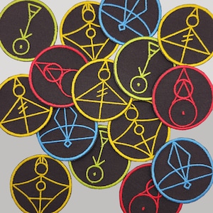 May include: A collection of black circular patches embroidered with colorful geometric designs. The patches feature yellow, blue, red, and green stitching. The designs include lines, circles, and geometric shapes. The patches are arranged on a light gray surface.