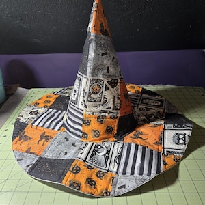 May include: A quilted witch hat featuring a patchwork design of orange, black, and white fabric with patterns of cats, skulls, and occult symbols. The hat sits on a green cutting mat with visible inch markings, showing a diameter of 16 inches.