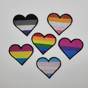 May include: Six embroidered heart patches with black borders. Each heart features a different color scheme, including rainbow, pink, blue, yellow, and black, representing various LGBTQ+ identities. These patches are ideal for customizing clothing or accessories.