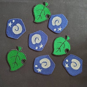 May include: A collection of embroidered patches featuring designs from the Animal Crossing video game. The patches include green leaves and blue, hexagonal shapes with a spiral and stars.