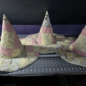 May include: Three quilted witch hats with wide brims. The hats are made of patchwork fabric in shades of pink, yellow, and green. Each hat has a tall, pointed crown. Numbers 1, 2, and 3 are visible on the crowns.