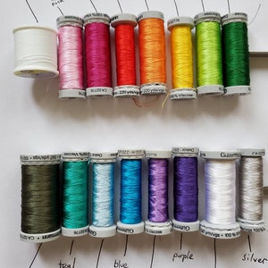 Puede incluir: Una colecci&oacute;n de coloridas bobinas de hilo, que incluyen tonos de rosa, rojo, naranja, amarillo, verde, azul, morado, blanco y plateado. Las bobinas est&aacute;n etiquetadas con nombres de colores y tipo de hilo, perfectas para proyectos de costura y manualidades.