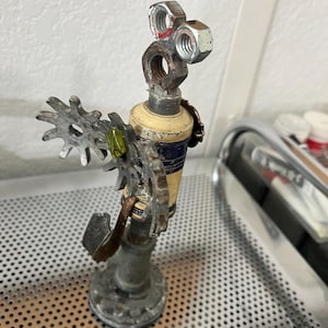 Könnte beinhalten: Eine Skulptur aus wiederverwendeten Metallteilen mit einem zentralen zylindrischen Element in Creme und Blau, das von zwei Sechskantmuttern als Augen gekrönt wird und mit zahnradförmigen Metallstücken sowie einer kleinen grünen Perle verziert ist.