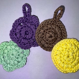 May include: Four crocheted dishcloths in different colors: green, purple, brown, and yellow. Each dishcloth has a flower-like design in the center.