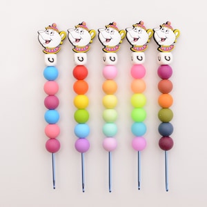 Puede incluir: Cinco collares de cuentas coloridas con un dije de tetera y una cuenta con la letra 'C' en la parte superior. Las cuentas están dispuestas en un patrón de arcoíris, comenzando con rojo y terminando con morado.