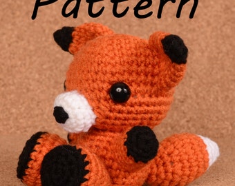 May include: A crocheted orange fox plush toy with black eyes and black accents on the ears, paws, and tail. The toy is sitting on a brown surface.