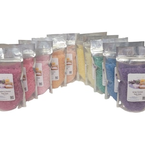 May include: A collection of ten clear stand-up pouches filled with colorful bath salts in shades of pink, orange, yellow, green, blue, and purple. Each pouch features a white rectangular label with text and a small image of soap and flowers.