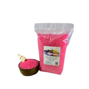 May include: A large bag of pink bath salts with a label that reads "Rose Petal Scented Bath Salts". The bag is sitting on a white surface next to a small wooden bowl filled with pink bath salts.