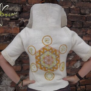 Op de afbeelding: Wit shirt met korte mouwen met een groot geborduurd geometrisch ontwerp in geel, oranje en roze. Het ontwerp bevat verschillende symbolen, waaronder een ster, een cirkel en een halve maan.