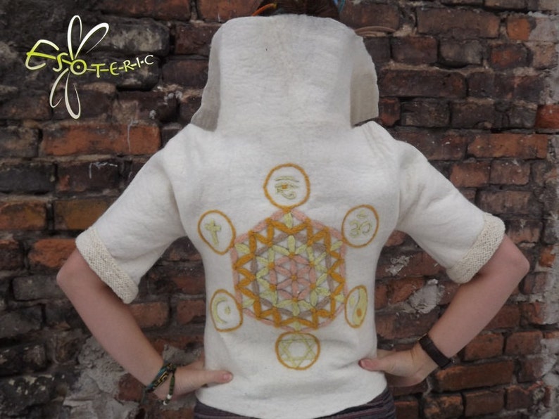 Op de afbeelding: Wit shirt met korte mouwen met een groot geborduurd geometrisch ontwerp in geel, oranje en roze. Het ontwerp bevat verschillende symbolen, waaronder een ster, een cirkel en een halve maan.