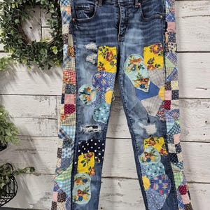 Op de afbeelding: Blauwe spijkerbroek met patchwork details. De jeans hebben kleurrijke stoffen patches met bloemen-, stippen- en ruitpatronen. De patches zijn op de voor- en zijkant van de jeans genaaid en geven een unieke, handgemaakte look. De jeans hangen aan een witte hanger.