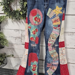 Op de afbeelding: Blauwe spijkerbroek met decoratieve patchwork-details. De spijkerbroek heeft bloemen- en geometrische patronen in rood, blauw en geel. De patchwork is op de voor- en zijkanten genaaid, wat een unieke, handgemaakte uitstraling creëert.