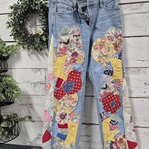 Op de afbeelding: Lichtblauwe spijkerbroek met patchwork bloemen- en geometrische ontwerpen in rood, geel en blauw. De jeans heeft een klassiek ontwerp met vijf zakken en een gerafelde zoom. Het patchwork heeft verschillende bloempatronen en vormen.