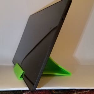 May include: A black tablet computer is propped up on a bright green stand. The stand is shaped like a triangle with a rounded edge.
