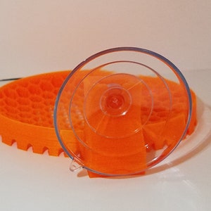 May include: A clear suction cup with a textured orange base. The suction cup is designed to hold a small object, such as a phone or a tablet.