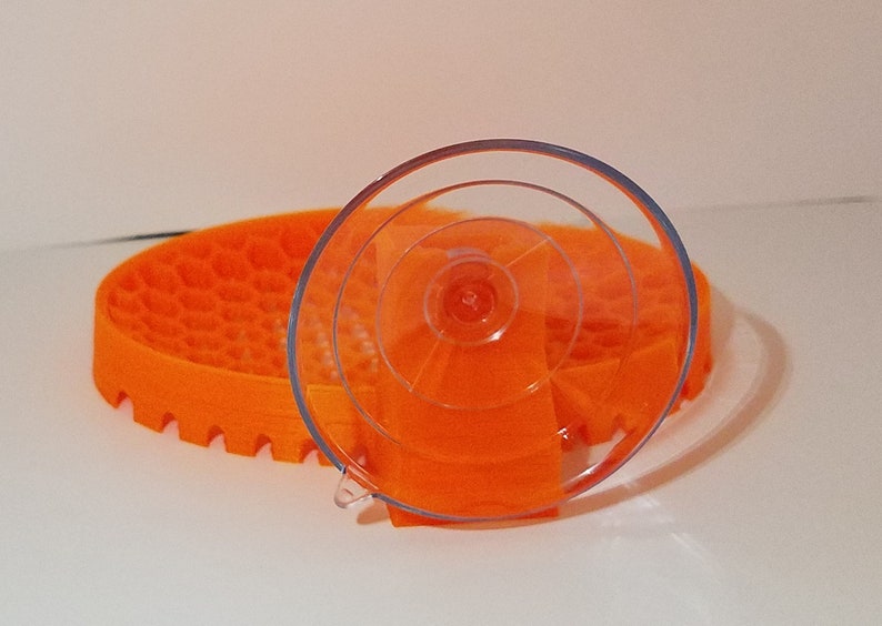 May include: A clear suction cup with a textured orange base. The suction cup is designed to hold a small object, such as a phone or a tablet.