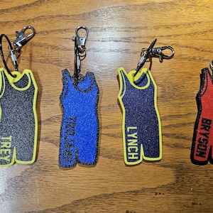 May include: Four wrestling singlets keychains with yellow trim. The singlets are black, blue, navy blue, and red. The names "Trey", "Trojans", "Lynch", and "Bryson" are printed on the front of each singlet.
