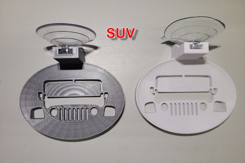 May include: Two 3D printed car-shaped suction cup holders. One is gray and the other is white. Both have a cutout of a Jeep SUV and a suction cup on top. The word "SUV" is printed in red on the image.